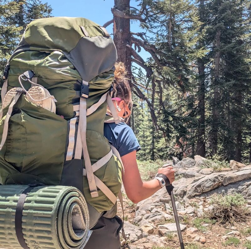 A woman is hiking in a forest. She is wearing a large backpack and using a hiking stick. The forest is dense with trees, and the ground is rocky. The sky is blue and clear.
